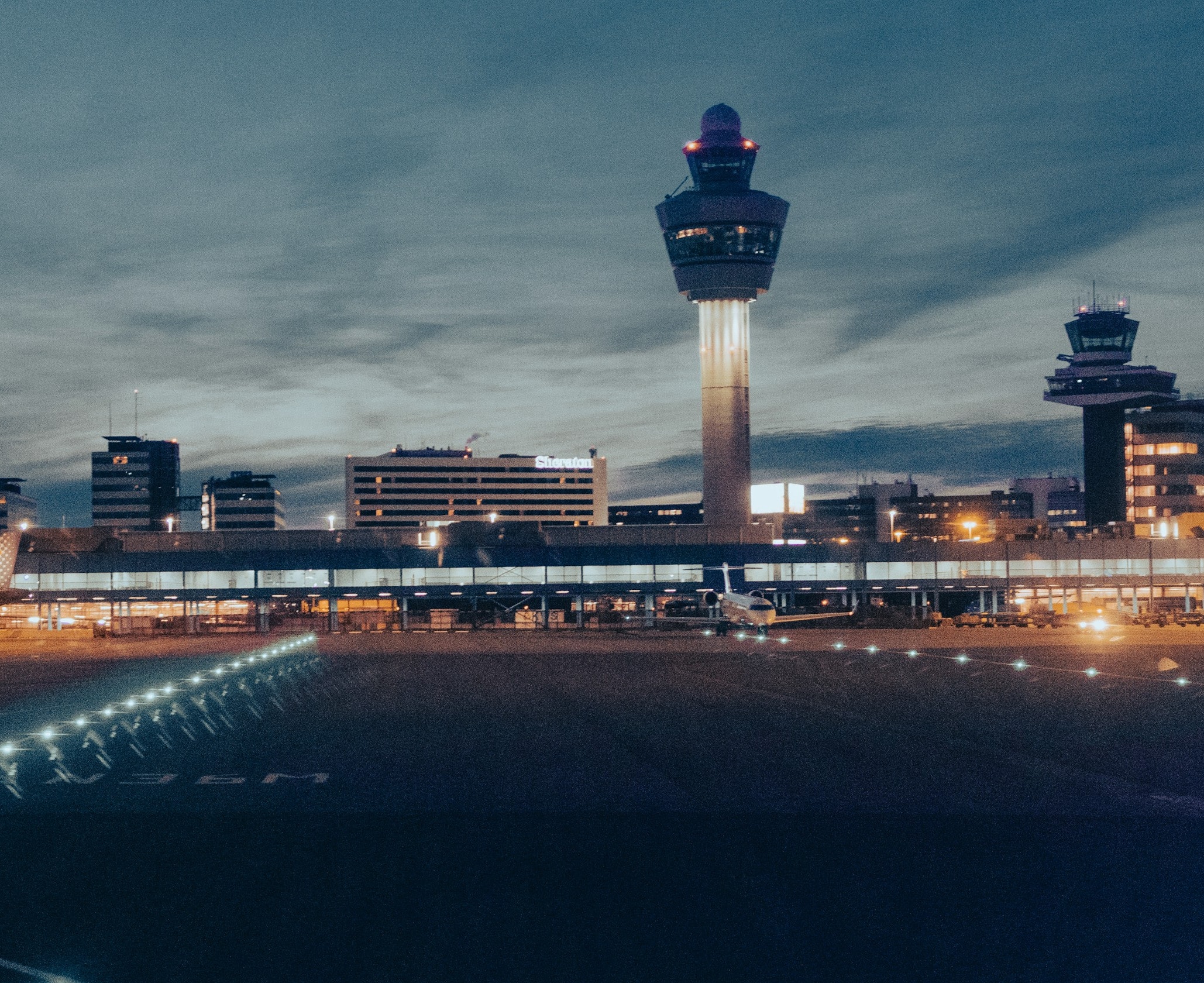 An airport terminal and apron at dusk, with taxiway lights in the foreground